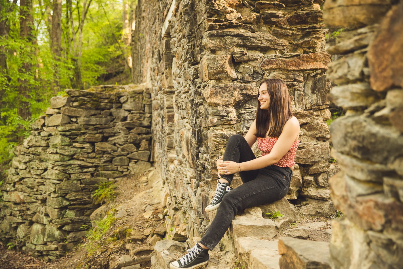 Smiling high school senior wearing black jeans, Converse, and a red tank top while sitting in old paper mill ruins and hugging her knee