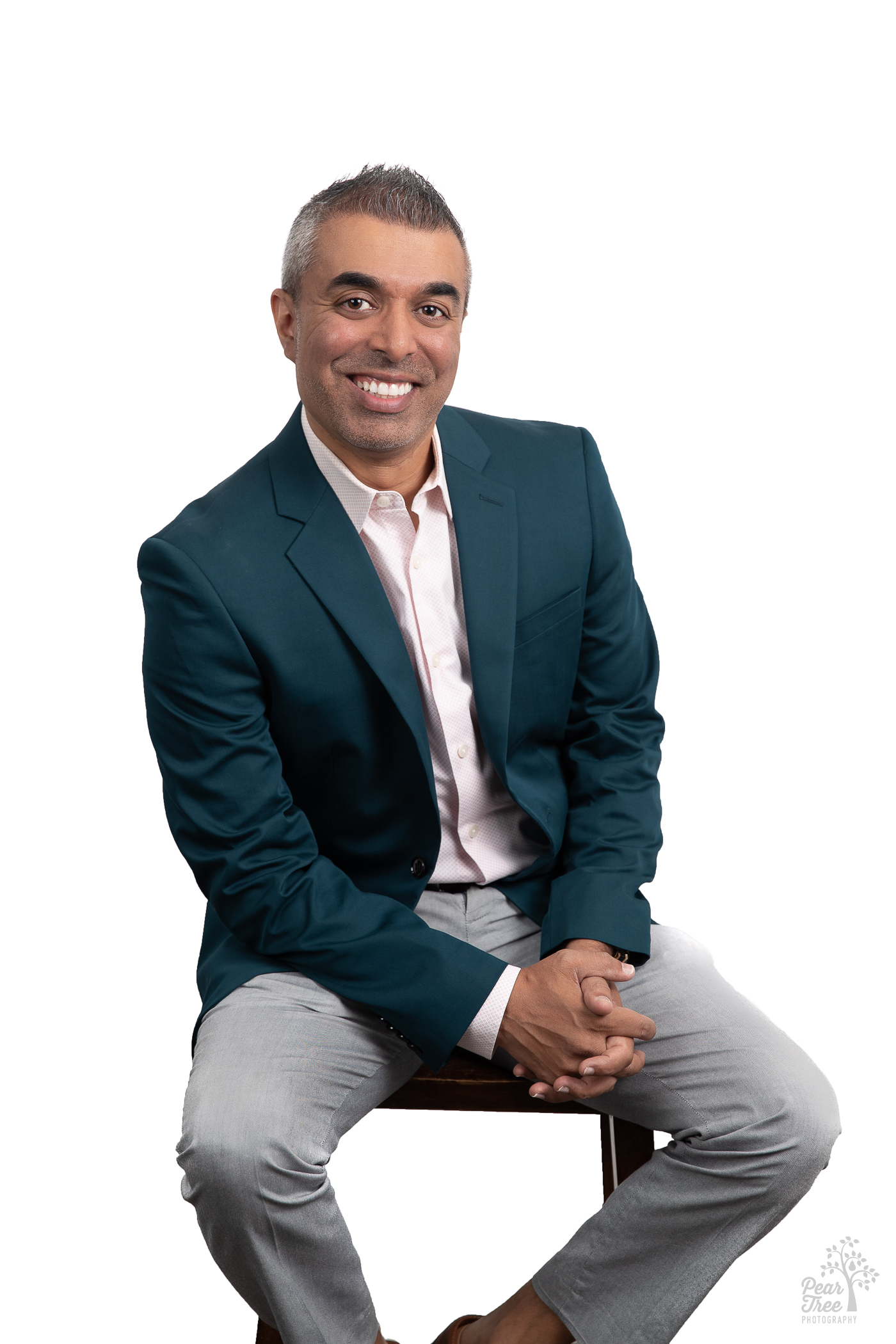 Engaging Indian American Realtor Minesh Patel sitting on a stool and smiling for realtor professional headshots. He's wearing grey slacks, a teal blazer, and light pink button down shirt.