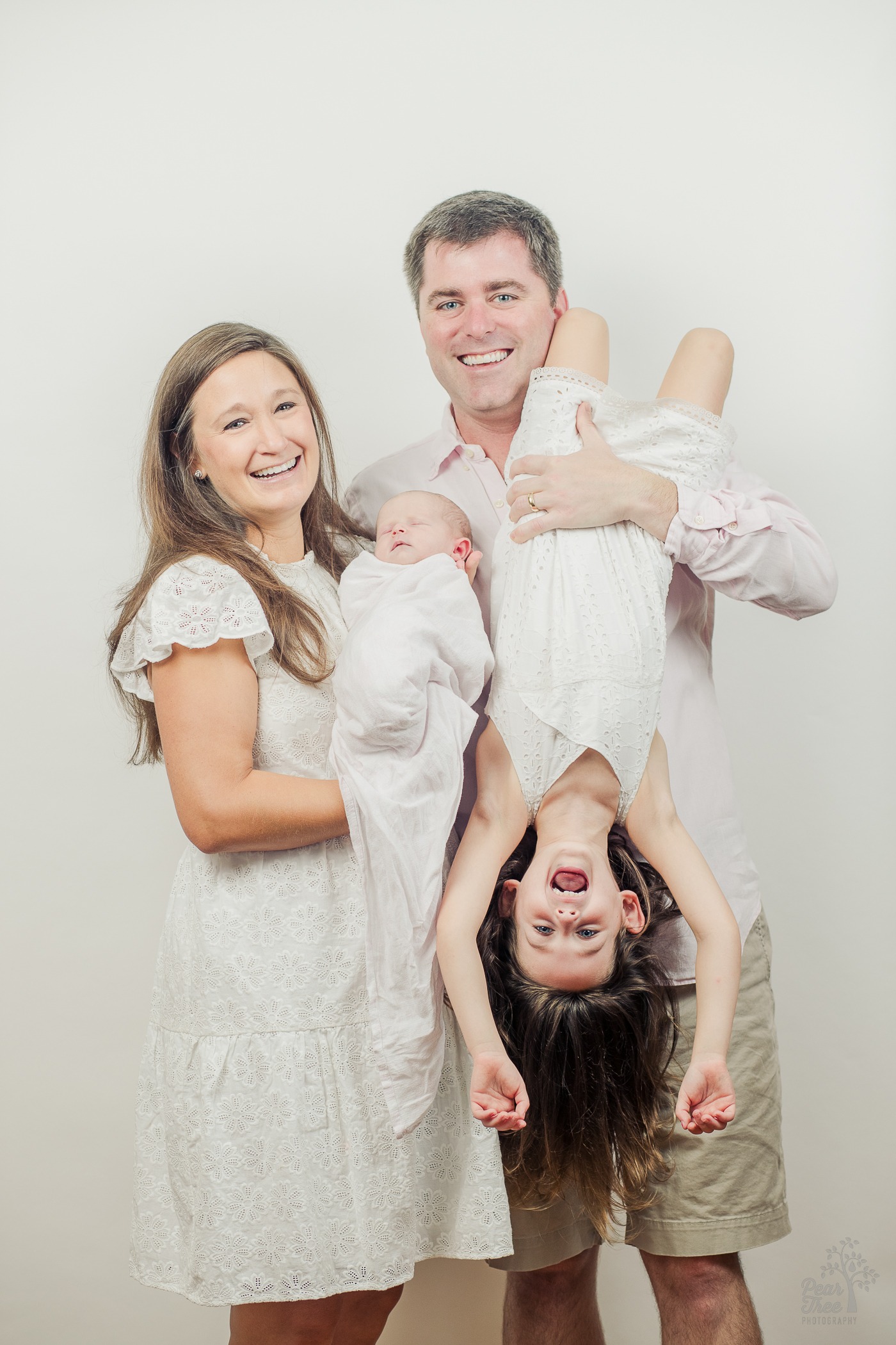 Mom and Dad holding their older and baby daughters. The older daughter is hanging upside down and laughing