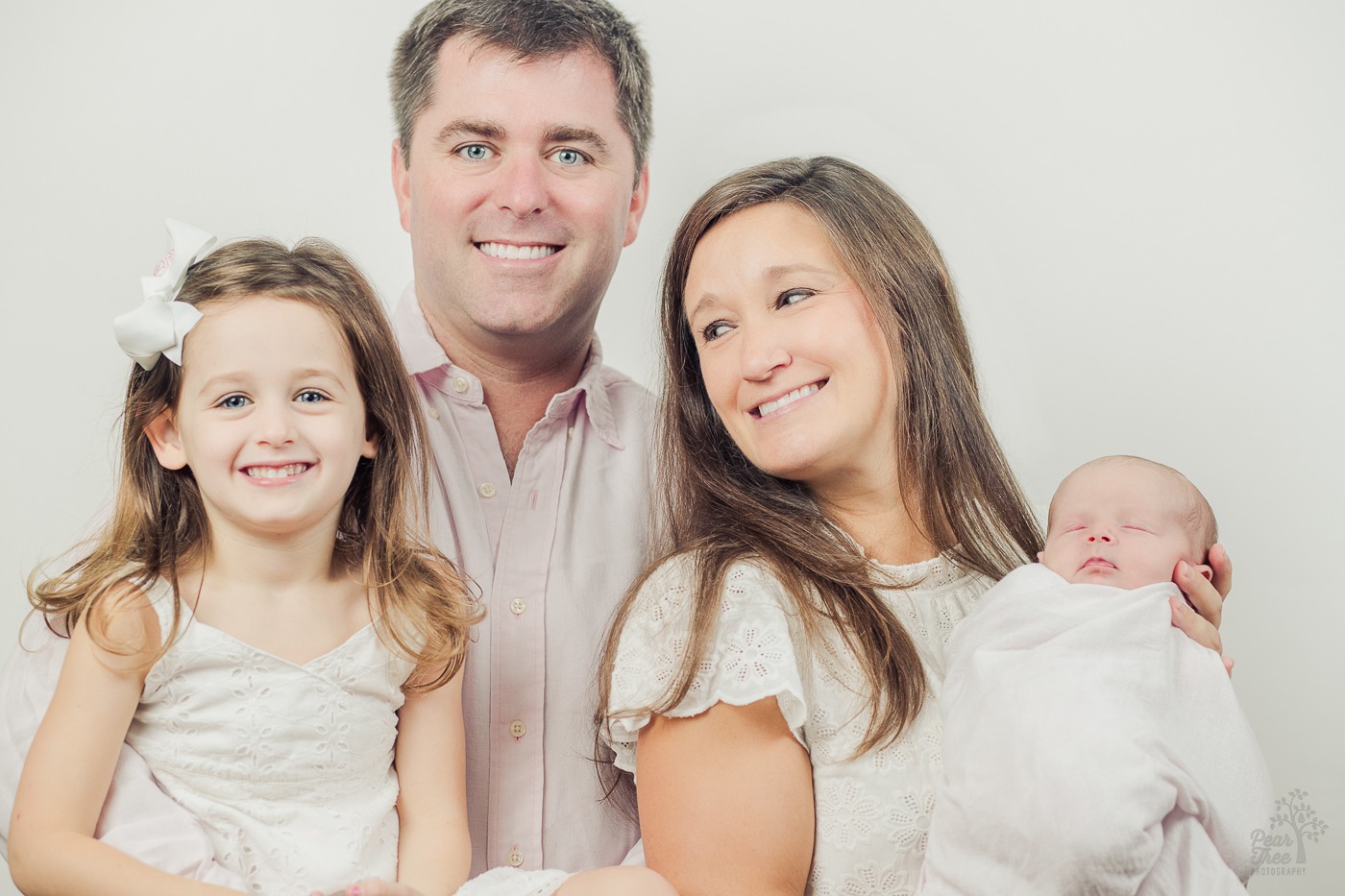 Dad and older daughter smiling at the camera. Mom is smiling at the older daughter while holding the baby for newborn photographs