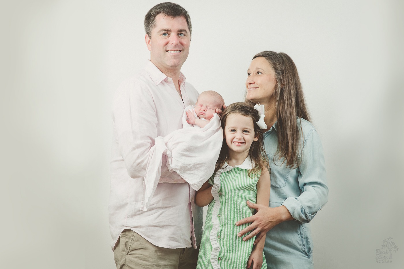 Dad and older daughter looking at the camera and smiling. Mom has her hand on her older daughter and smiling at her husband. Dad is holding their newborn daughter