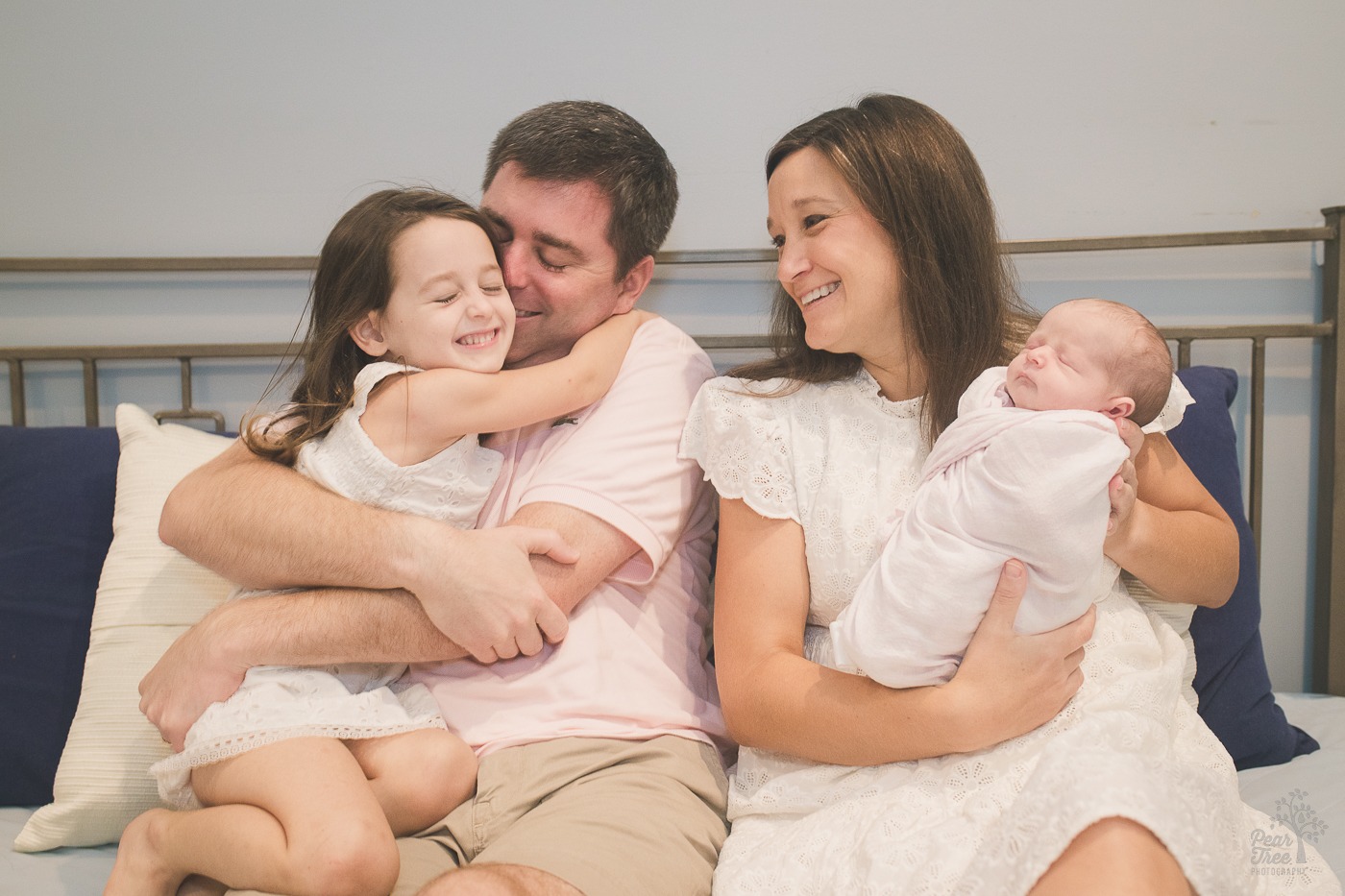 Father and daughter hugging while mom holds newborn baby and smiles at them on master bed