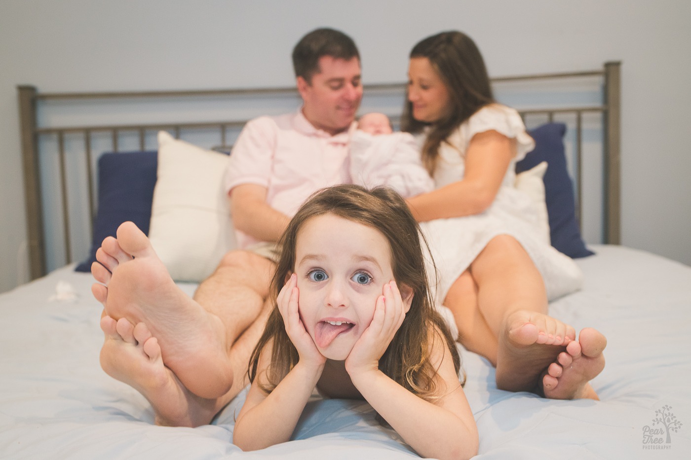 Cute little girl laying on her parents bed making a silly face while holding her face with her hands. Parents in the background smiling over their newborn