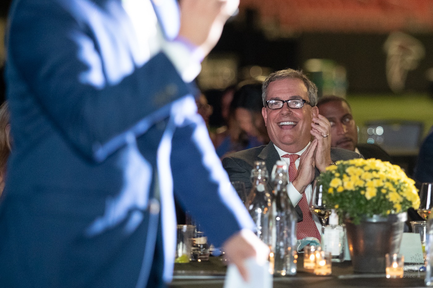 Sandy Douglas sitting at a table clapping his hands in joy. The auctioneer helps get folks excited to win auctions at the Night of Broadway Stars event for Covenant House Georgia