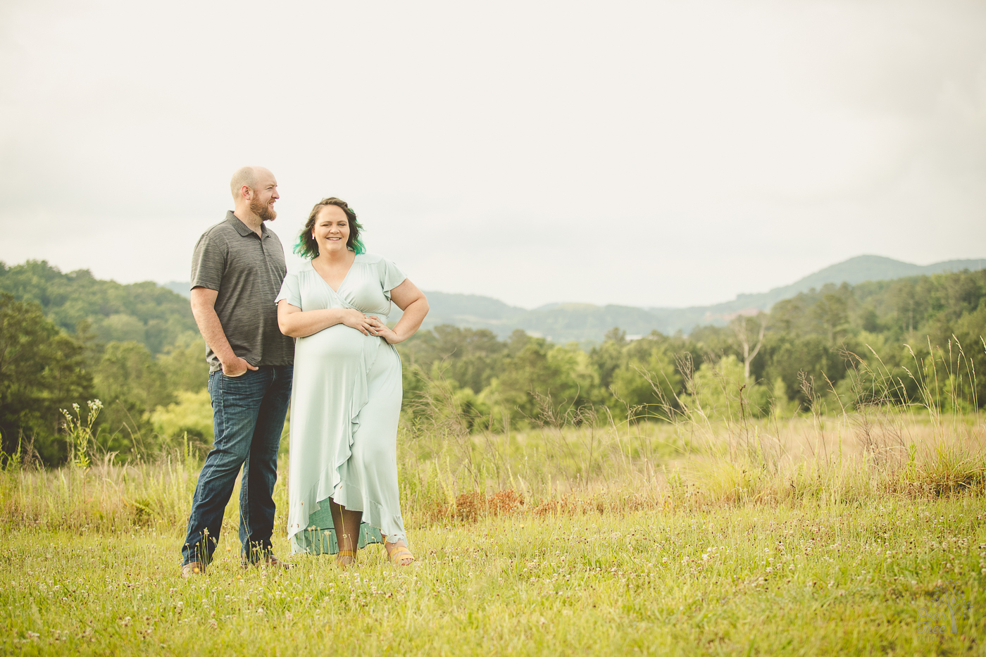 Beautiful pregnant woman standing in a long dress and hands over her big belly for maternity photographs. Her husband is standing next to her looking into the distance with his hands in his pockets.