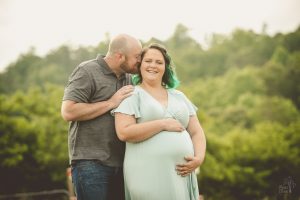 Pregnant mom holding her belly in a green dress. Her husband has her hand on her shoulder and is pushing his nose into her ear