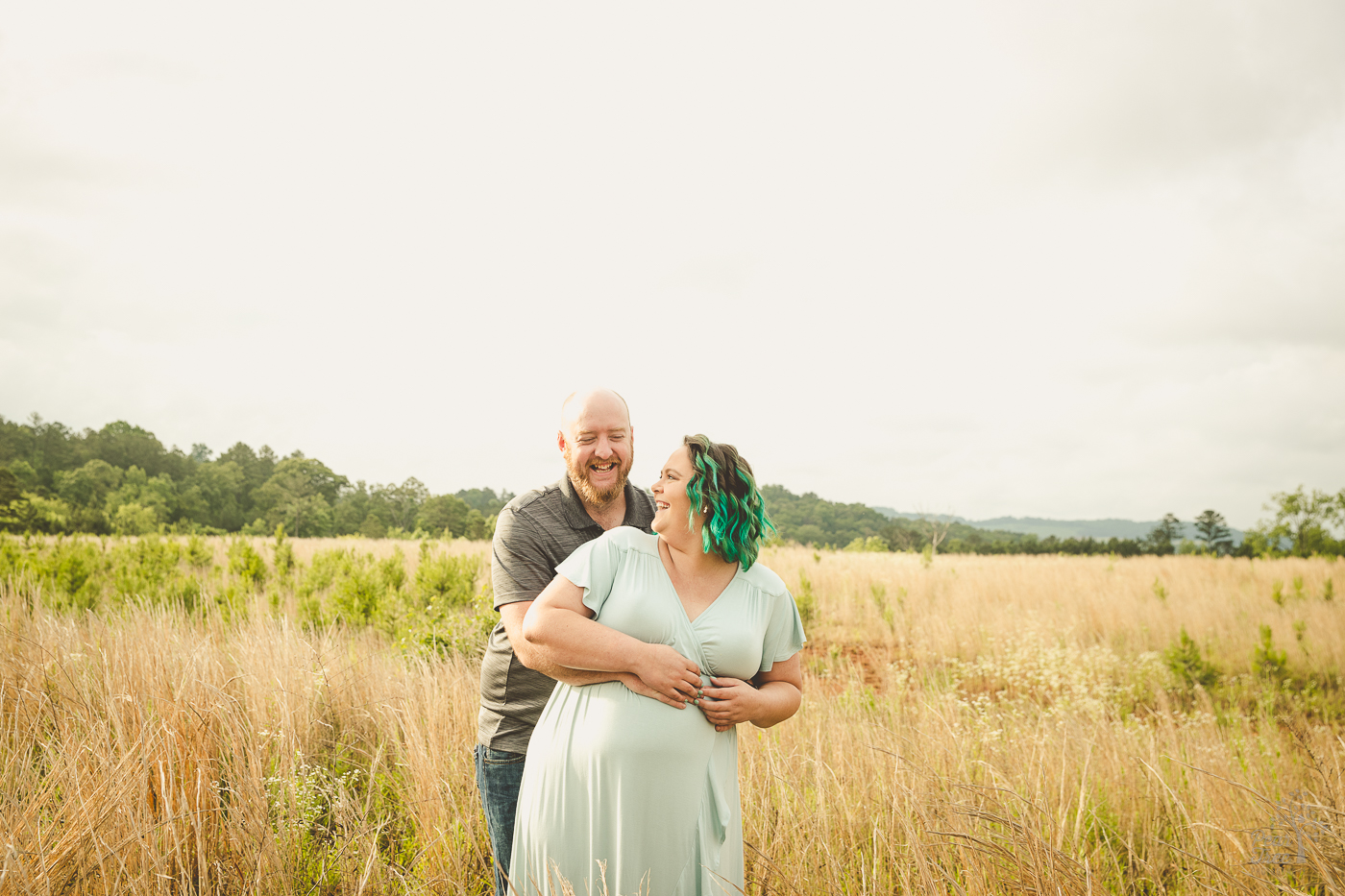 Pregnant couple holding each other and laughing in a field of tall grasses