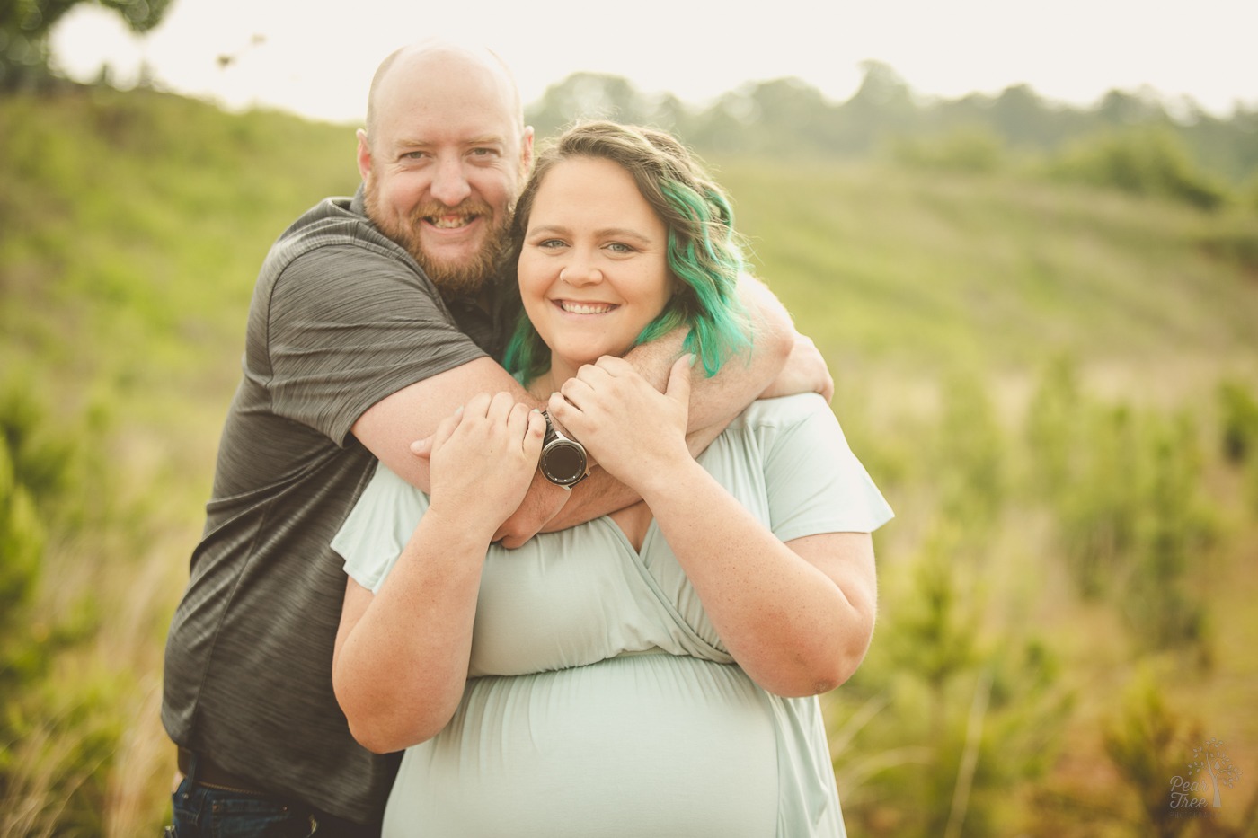 Husband wrapping his arms around his pregnant wife's neck and upper body. Pregnant mom is holding onto her husband's arm and smiling