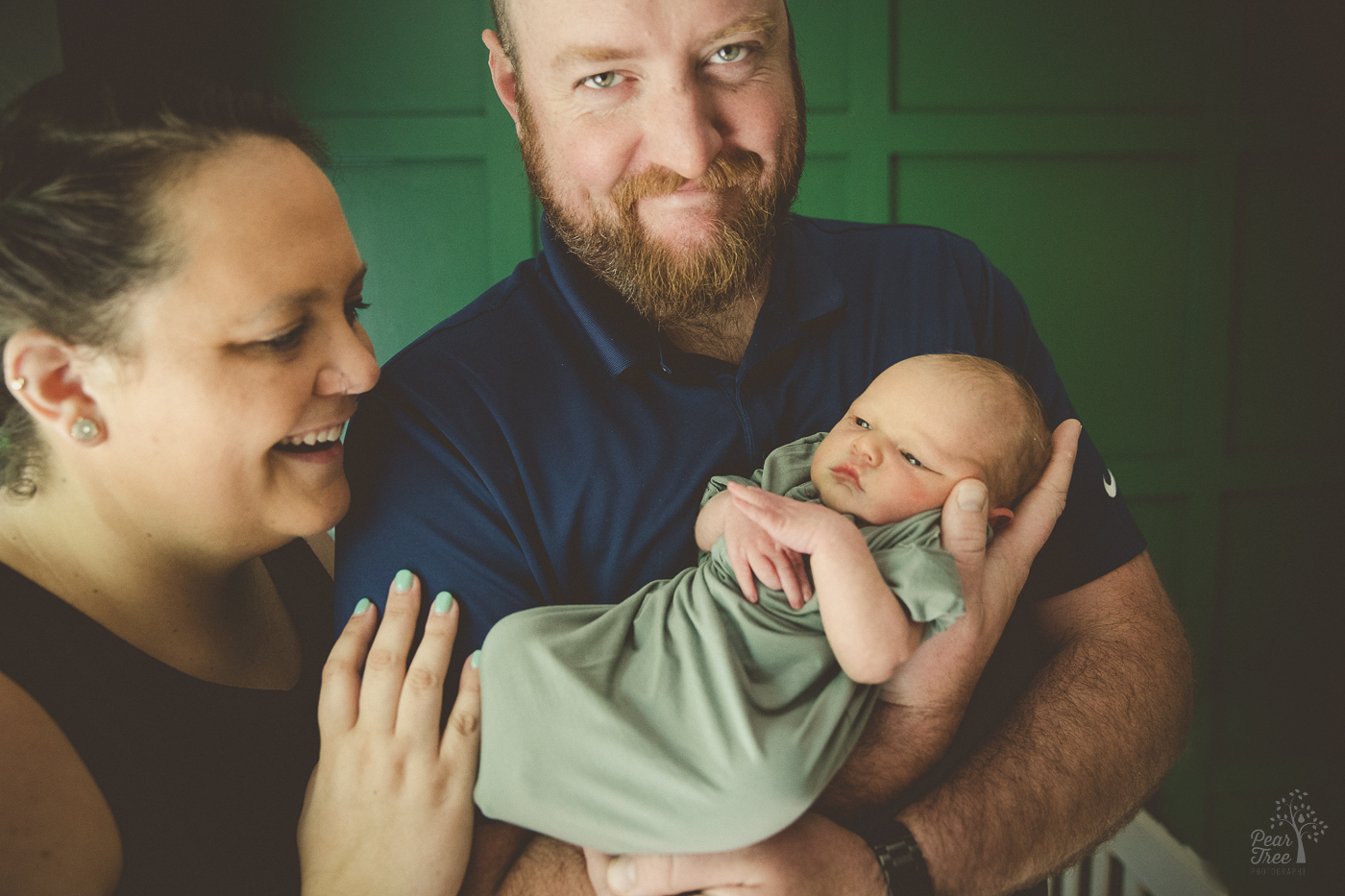 Proud dad holding his newborn son with mom smiling and her hand on his arm during their newborn photography session