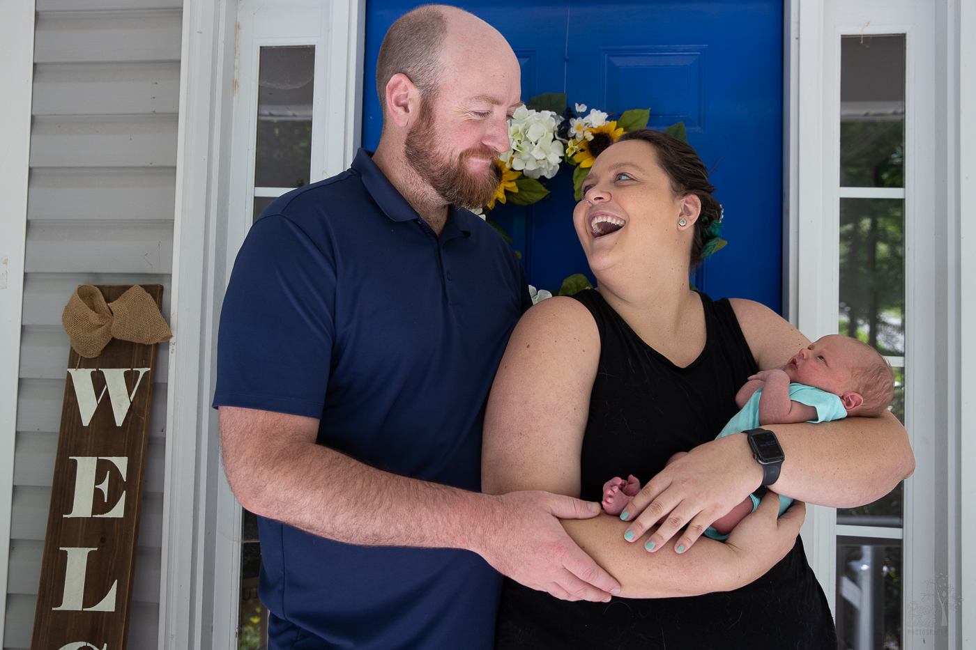 Parents standing in front of their blue front door holding their newborn son and laughing