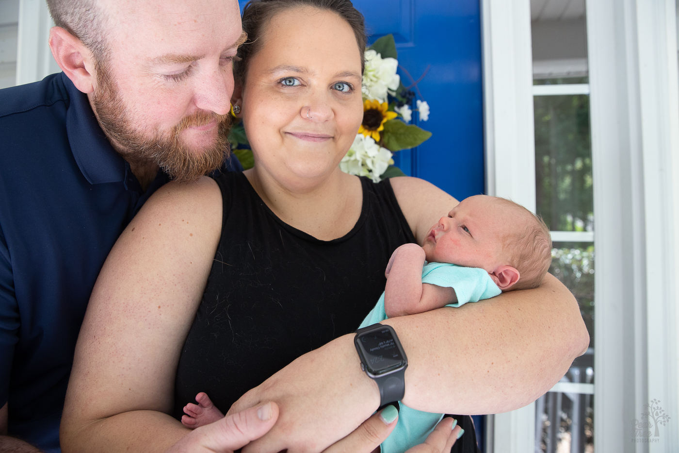 Mom and dad holding their infant son and smiling in front of their front door at the end of their newborn photography session