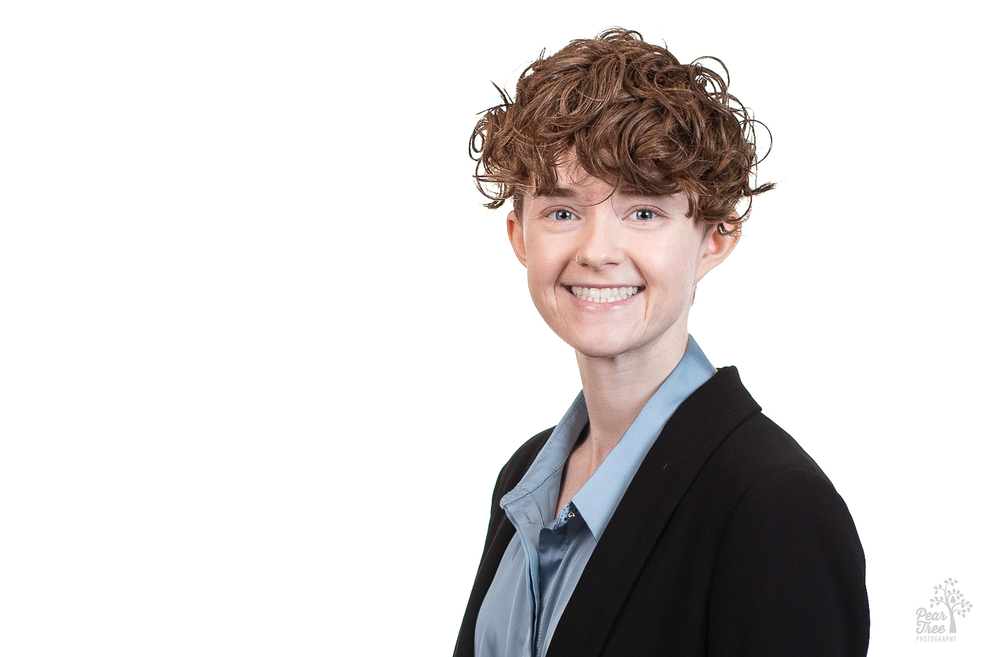 Caucasian woman with short, curly red hair is wearing a blue blouse and black blazer while smiling big