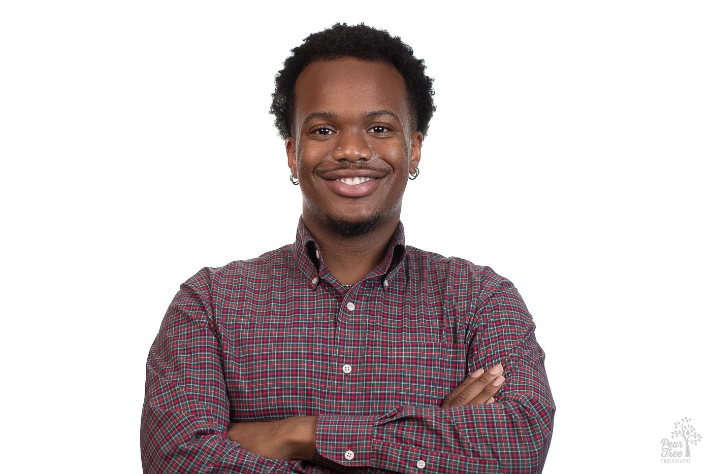 Attractive African American man wearing earrings and crossing his arms in a plaid button down shirt while smiling for a professional headshot in front of a white background
