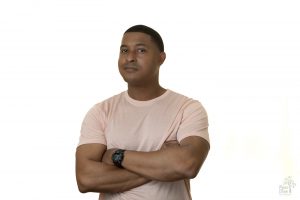 Professional headshot of an attractive African American male. He's smirking with his arms crossed while wearing a black watch, pink shirt and standing in front of a white background.