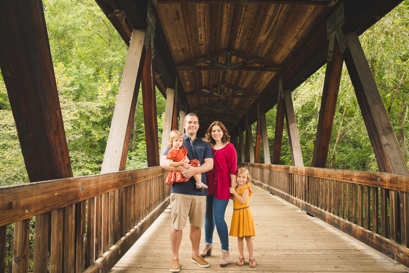 Beautiful family with two little girls wearing dresses standing together holding each other in the Roswell Mill covered bridge and smiling for family photographs