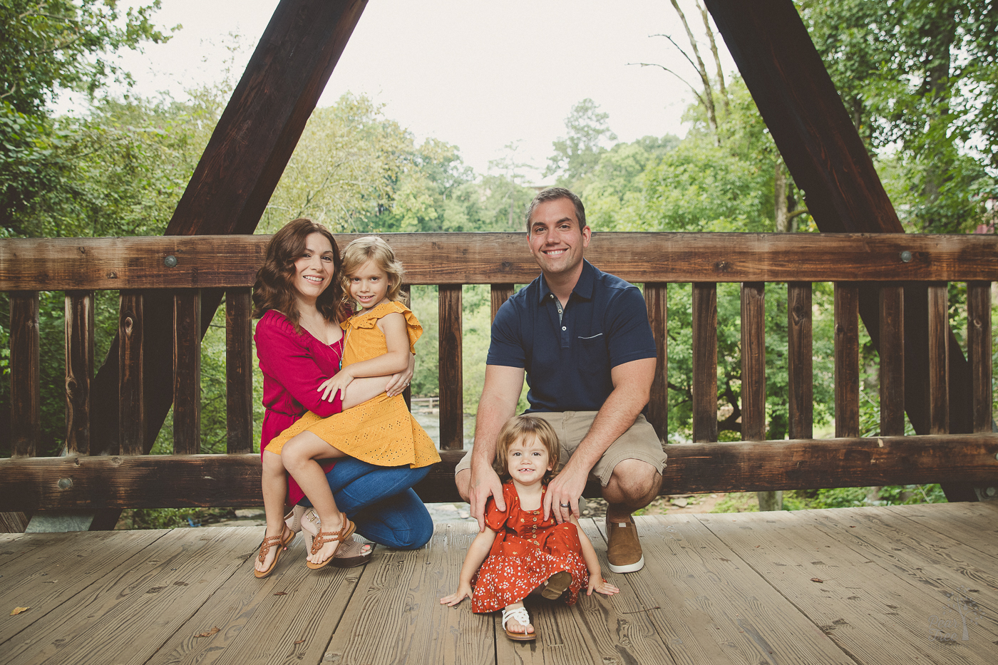 Two parents taking a knee on Roswell Mill covered bridge and holding their daughters while all smiling for family photographs