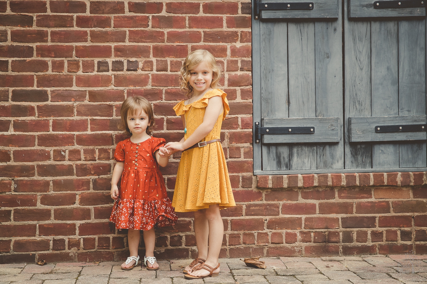 Two sisters holding hands in front of a brick building wearing dresses and smiling