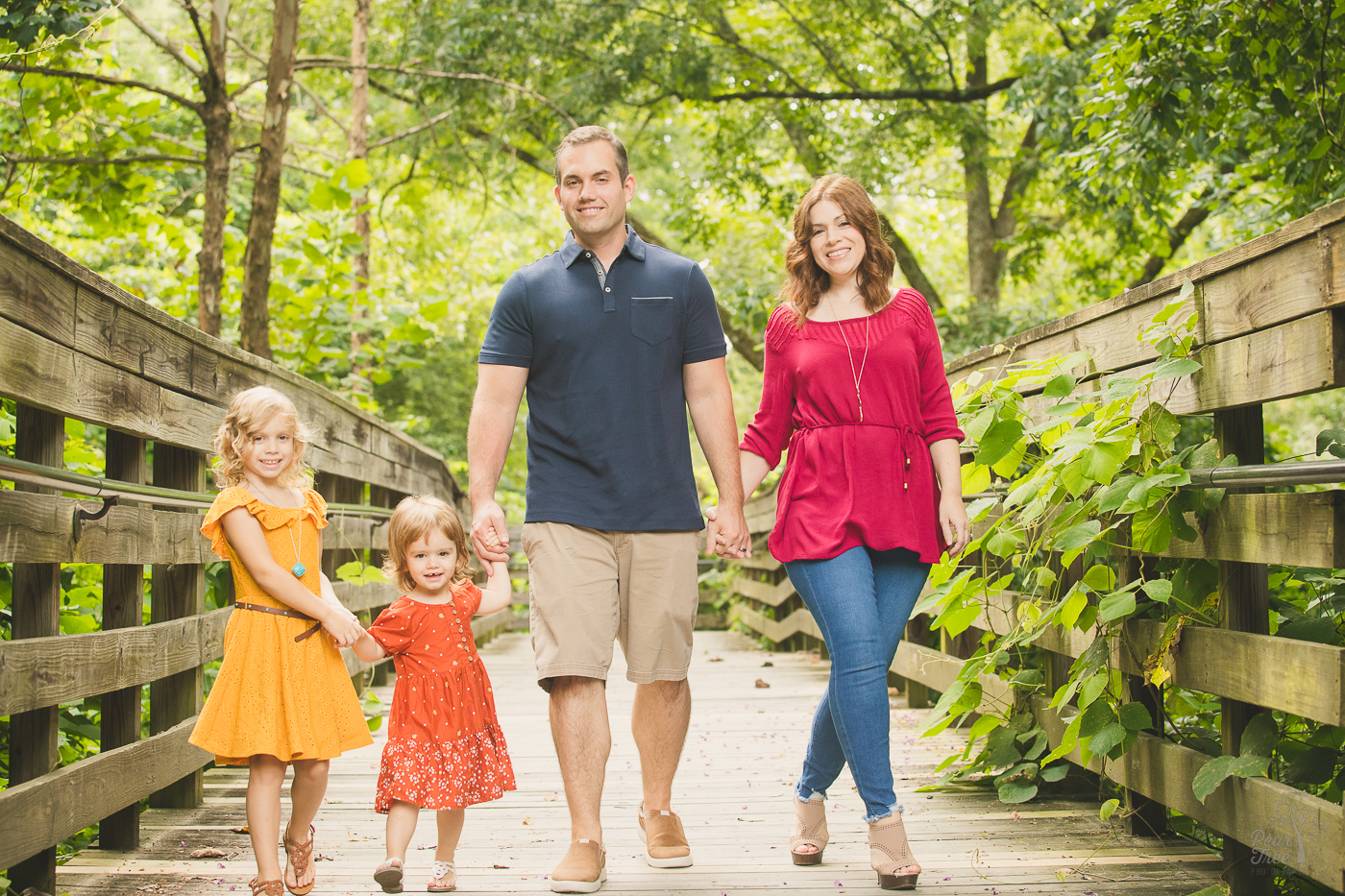 Mom and Dad holding hands while walking down the Roswell Mill boardwalk towards the camera with their two young daughters while smiling for family photographs and dressed fashionably