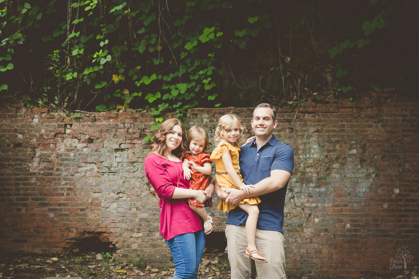 Mom and Dad holding their two young daughters in dresses on their hips and smiling in front of old mill ruins