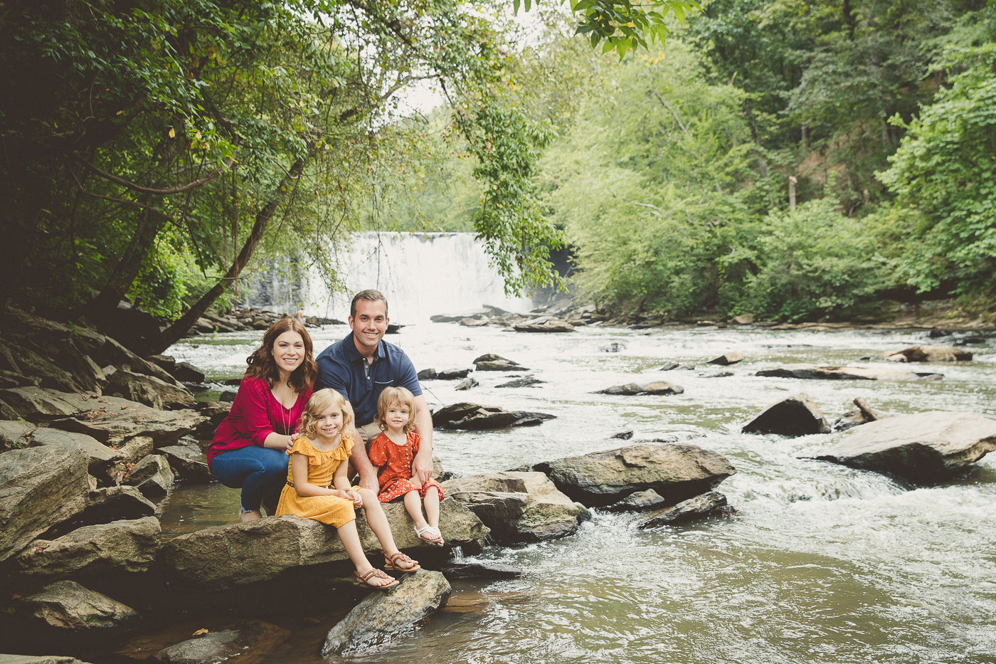 Mom and Dad squatting behind their two daughters sitting on rocks along the shoreline with the Roswell Mill dam waterfall behind them for family photographs