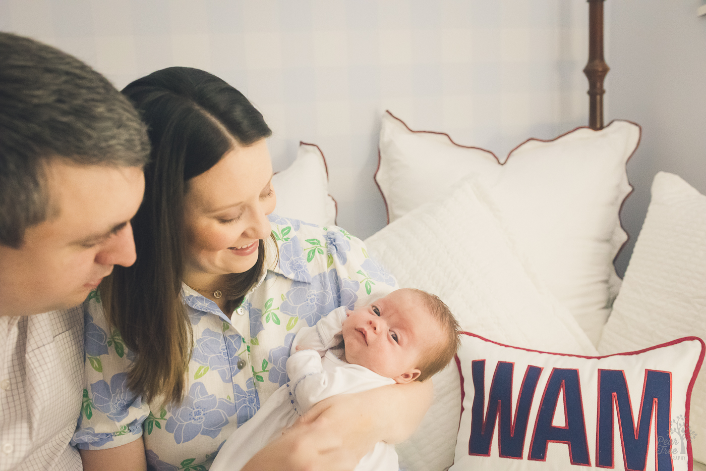 New parents sitting on a four poster bed holding their newborn son. One of the pillows on the bed has the baby's initials embroidered on it