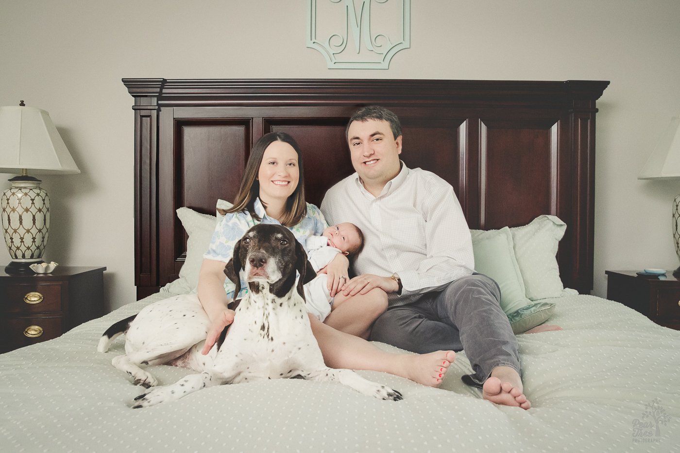 Parents with newborn son and family dog sitting on the master bed looking happily at the camera