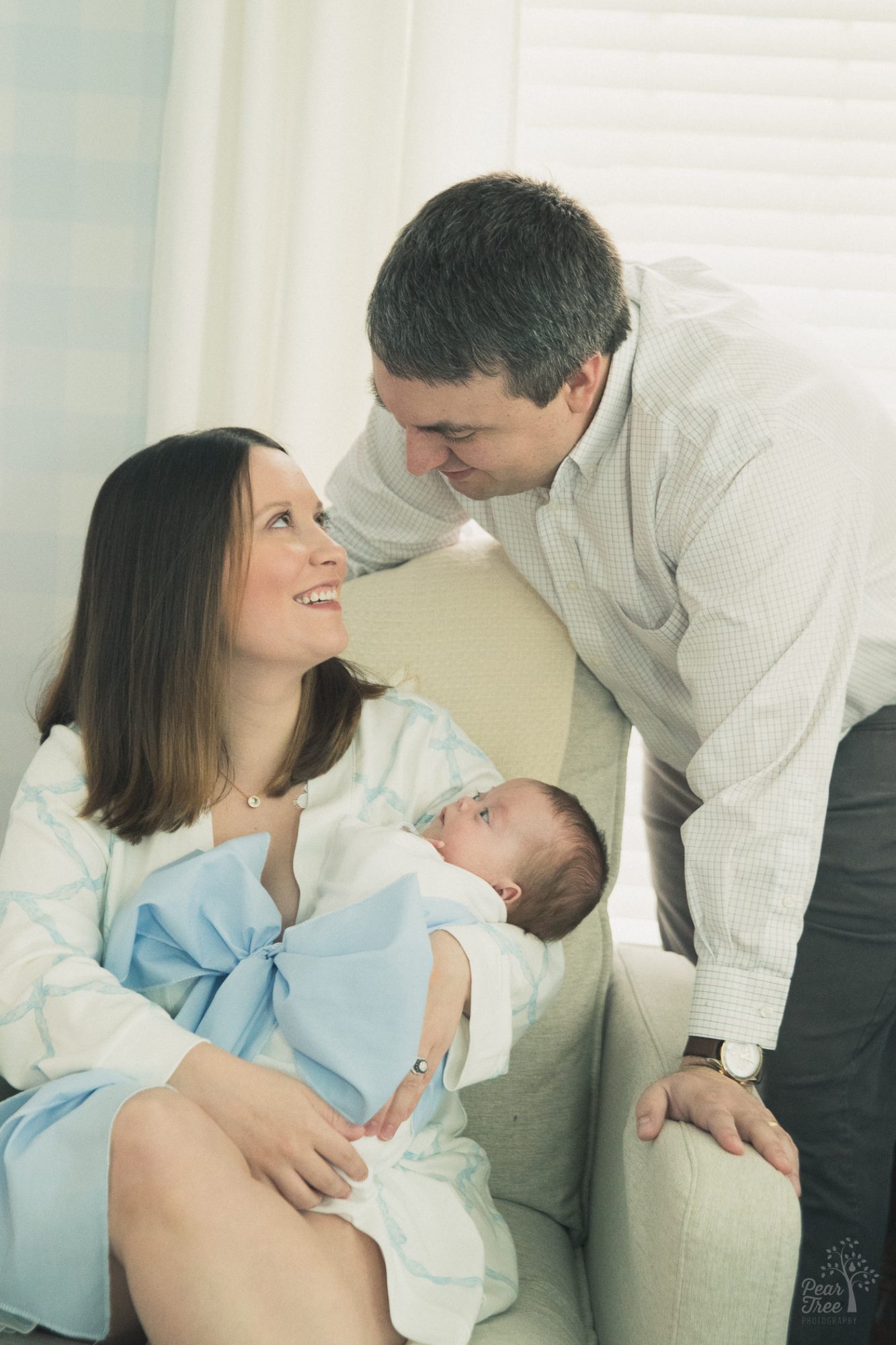 Mom sitting in chair smiling and looking up at dad while holding their newborn son wrapped in a blue bow