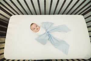 Newborn baby boy wrapped in a white blanket and big blue bow. Laying on his back looking up from the middle of his dark wood crib and white mattress