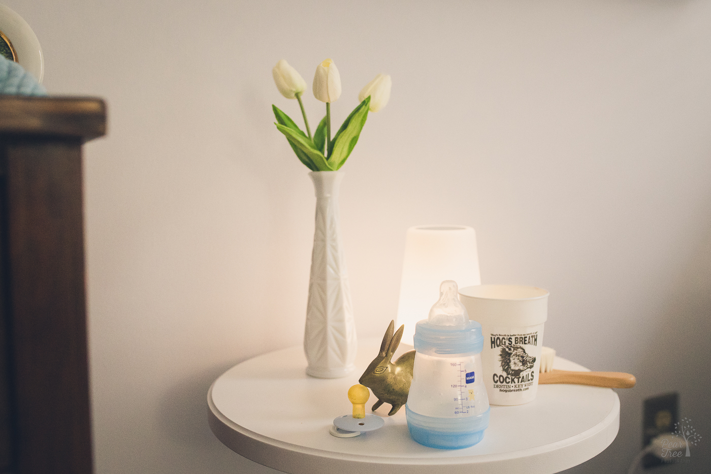 Small table in a nursery with a pacifier, bottle, gold rabbit, hairbrush, vase with roses, and a Hog's Breath Cocktails plastic cup