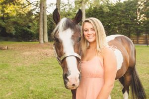 A blonde high school senior girl standing next to her horse and smiling