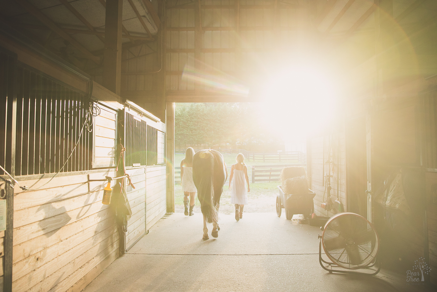 two girls in white sundresses walking their horse out of a stable at sunset