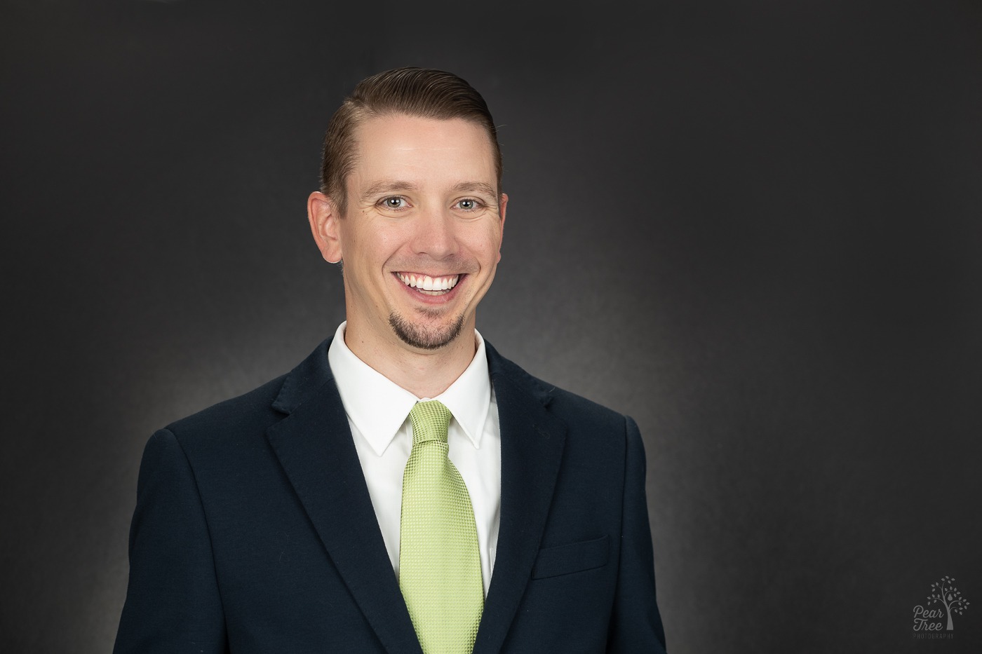 Professional headshot of a man smiling in front of a dark background. He's wearing a black suit, white shirt, and a green tie. Sporting a neatly trimmed goatee.