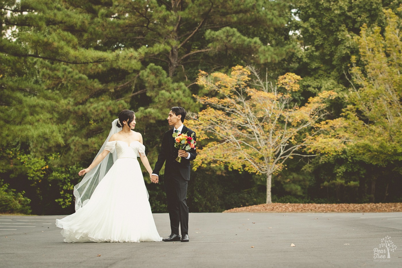 Newlywed Asian couple holding hands and smiling with fall leaves on trees behind them