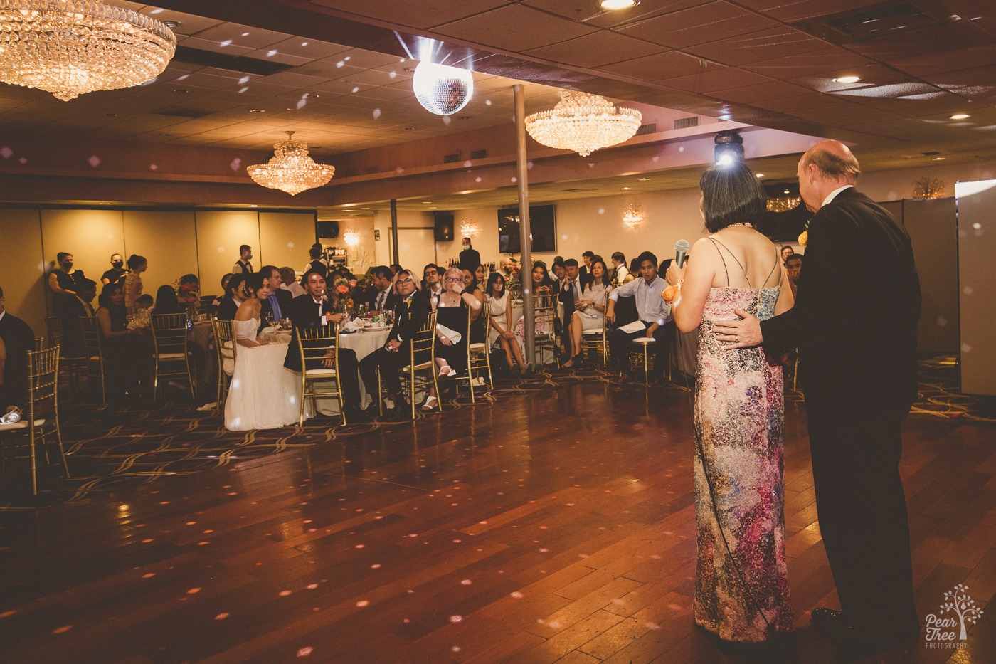 Bride's parents touching and giving a speech on stage while Asian American wedding couple sits at their table