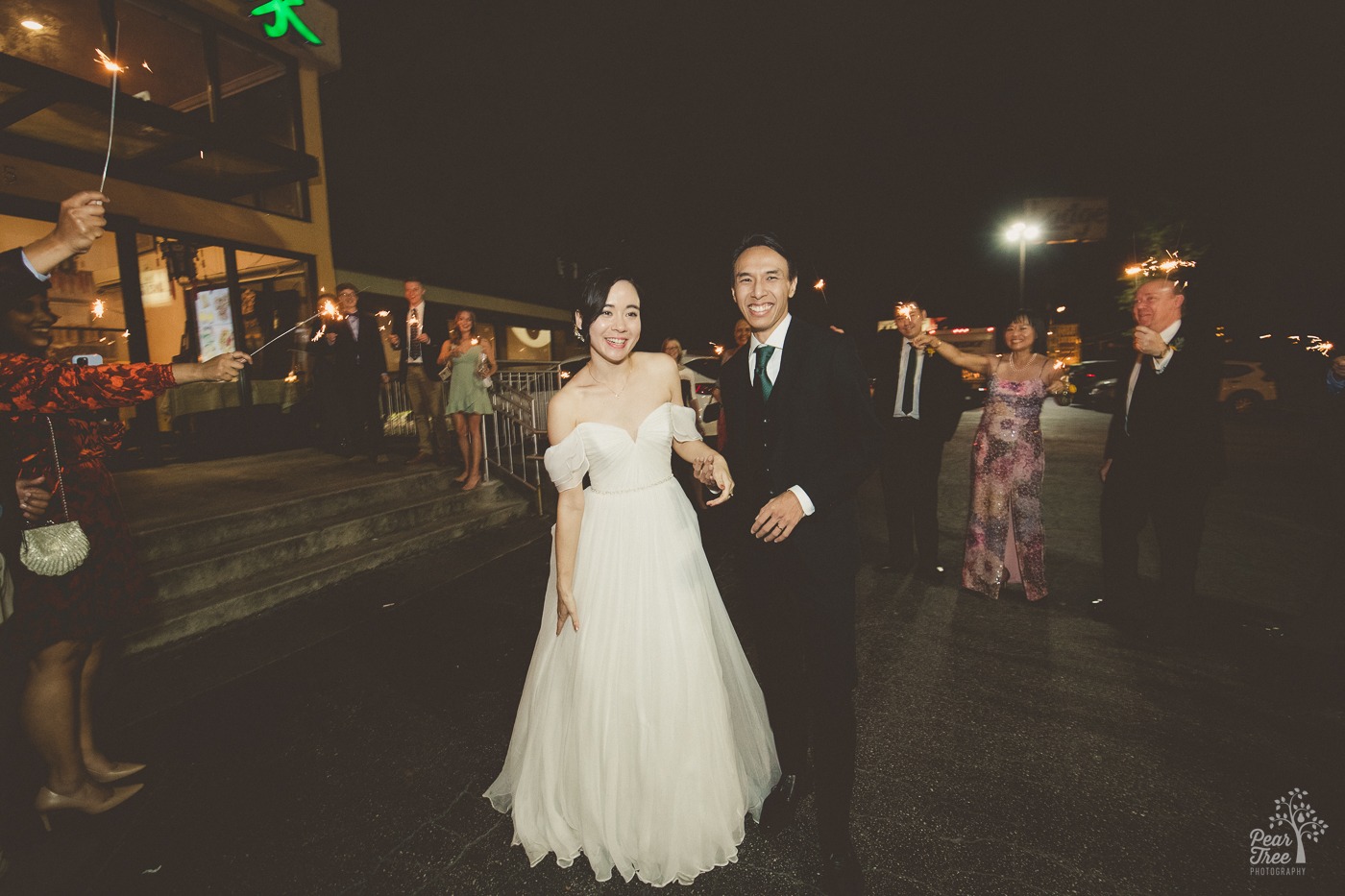 Smiling newlywed Asian couple standing in parking lot surrounded by happy family holding sparklers