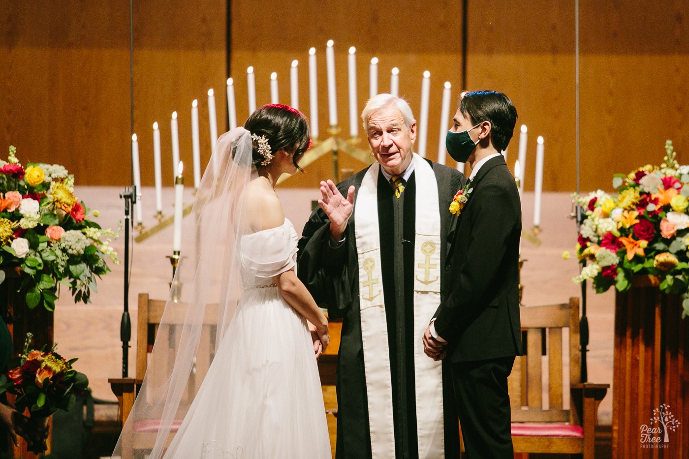 Officiant talking to bride on altar while groom looks on wearing a mask