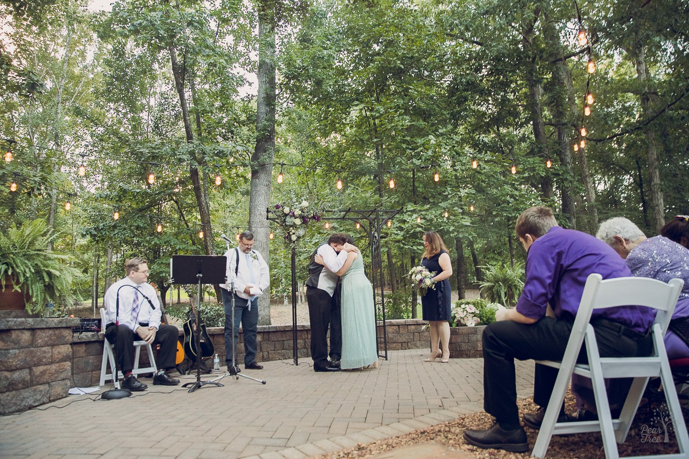 Bride and groom hugging while officiant prays and everyone bows their heads