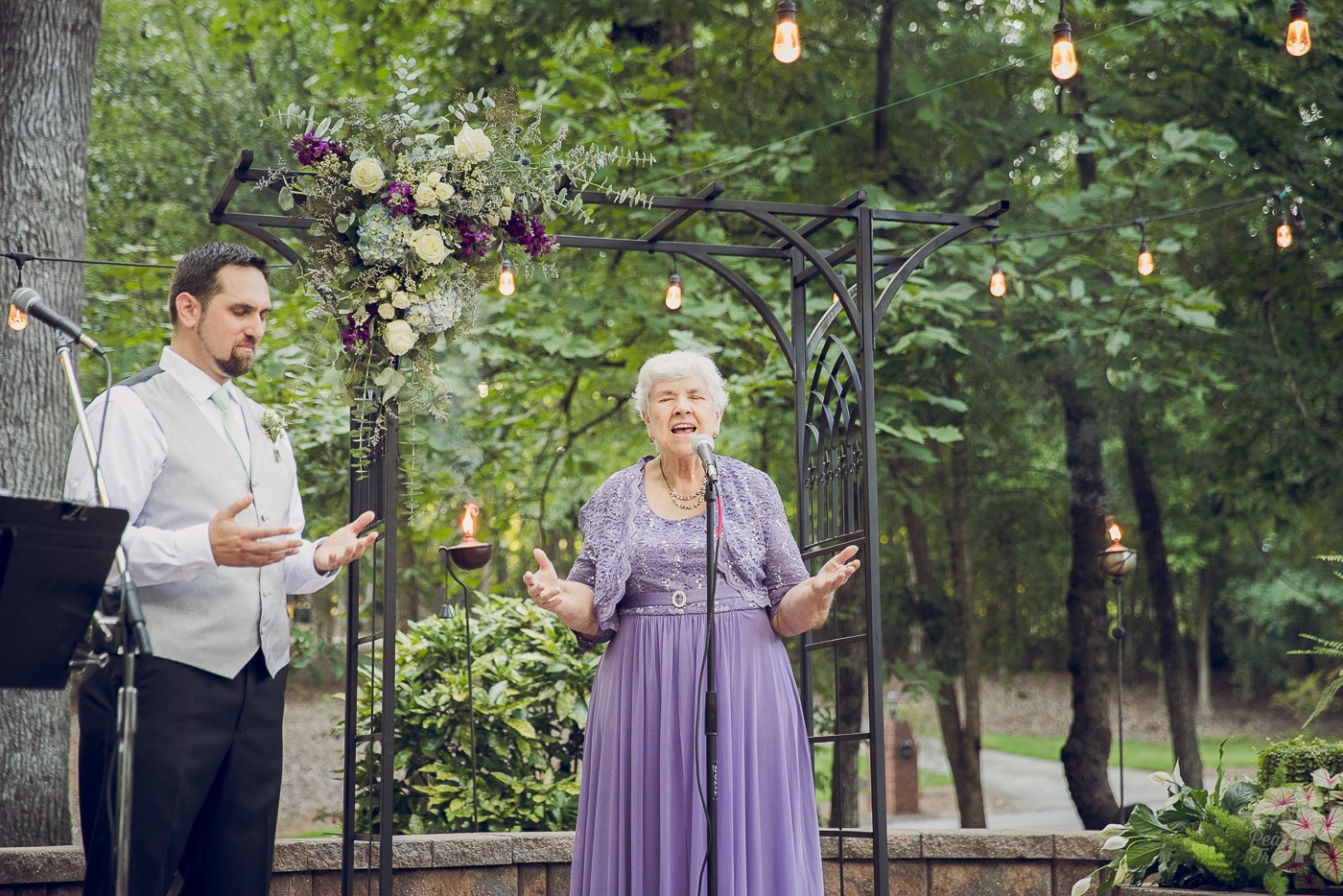Grandmother praying with hands outstretched at start of a wedding ceremony surrounded by green foliage and hanging lights