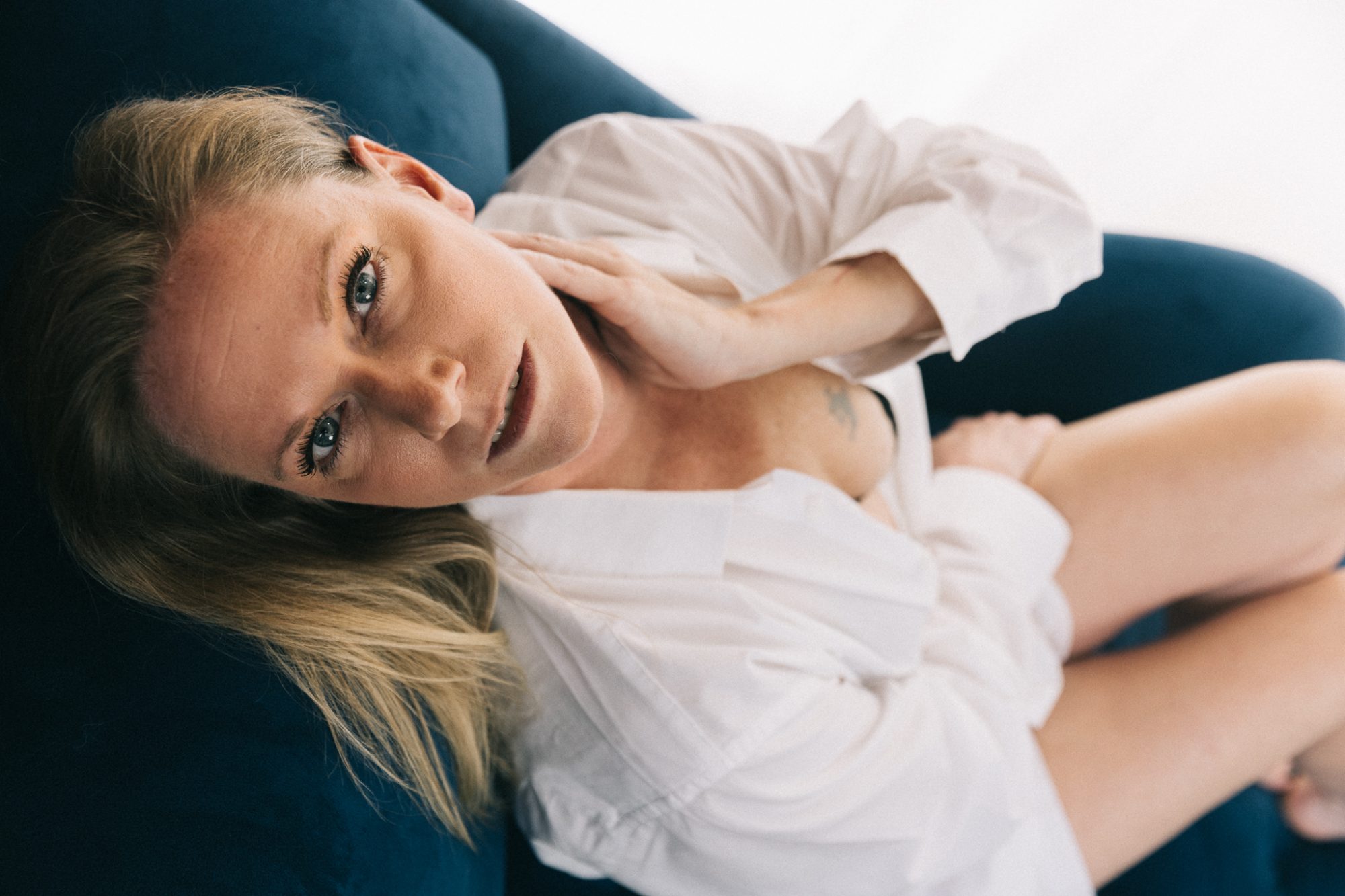Beautiful over 40 woman in men's white dress shirt and lingerie leaning back over a blue velvet chaise for boudoir photos