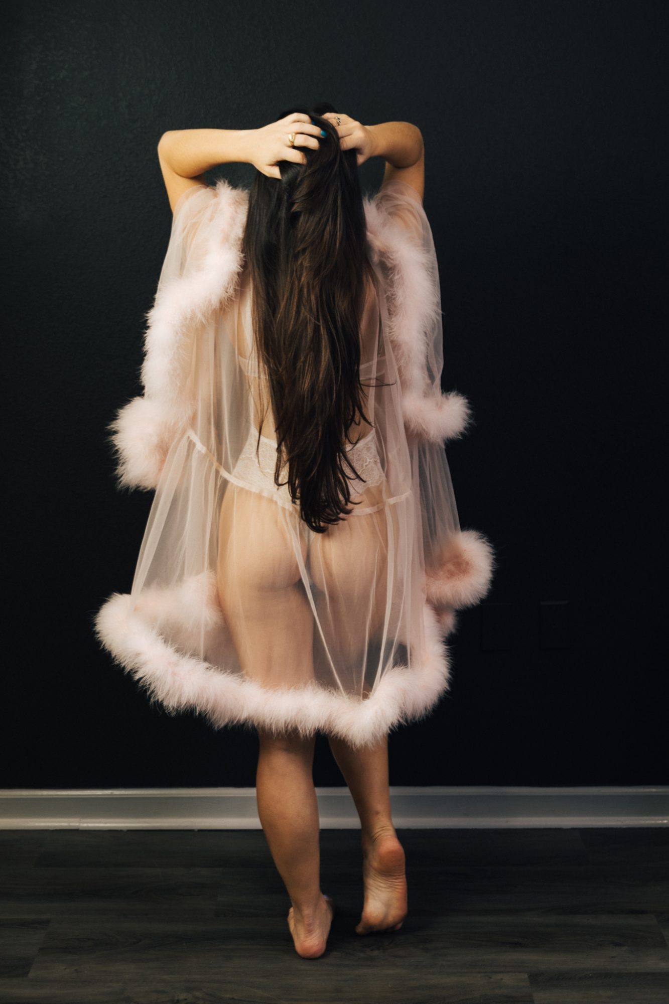 Back of a woman with hands on head wearing lacy lingerie under a sheer robe decorated in feathers for boudoir photos