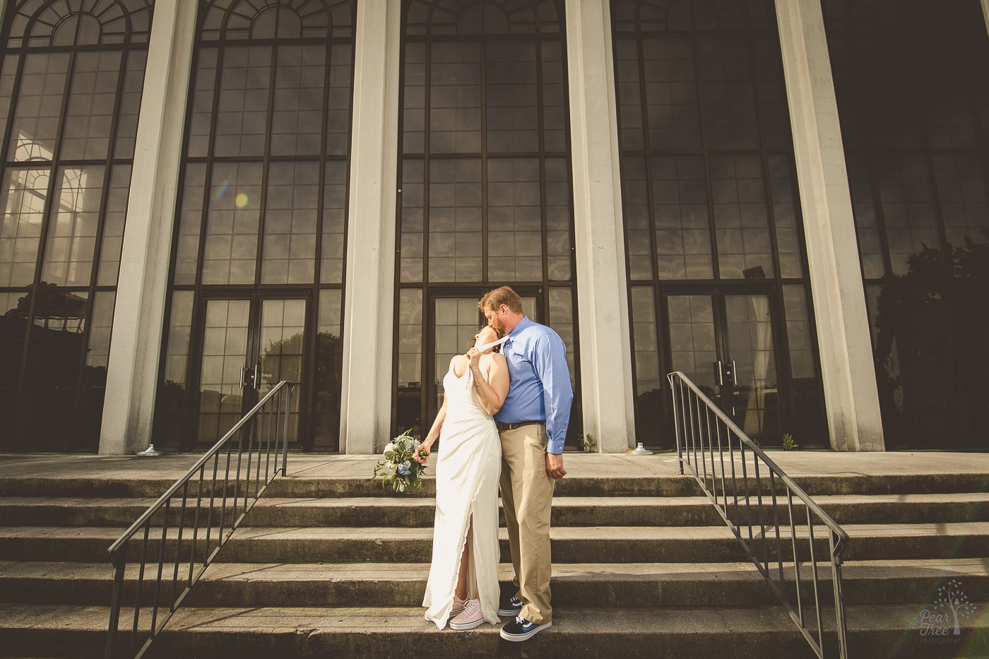 Bride holding grooms tie while she kisses him on steps in front of building with tall windows