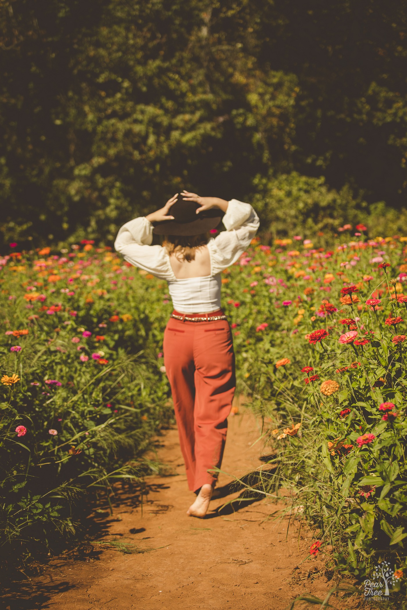 Creekview high school senior girl walking away barefoot through a flower field while holding her hat on her head