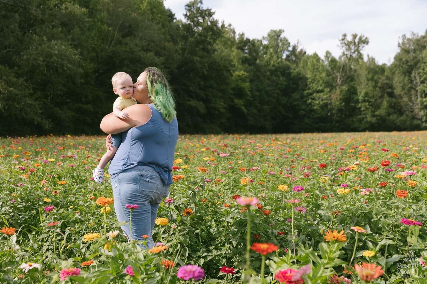 Mom with green hair holding her one year old son and kissing his cheek in a flower field