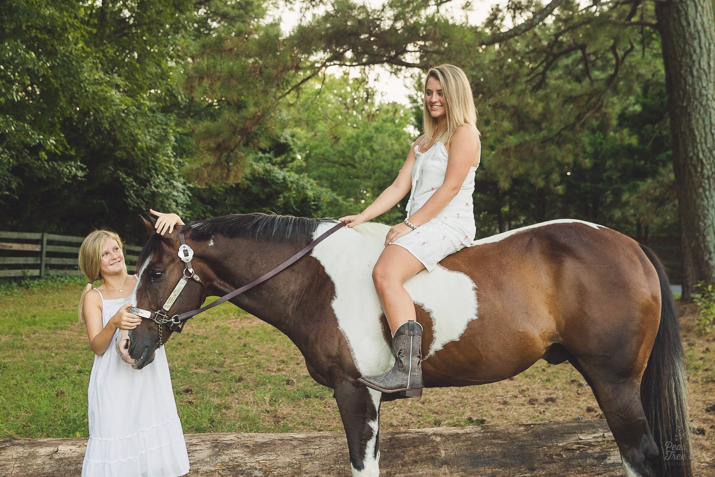 Younger sister fixing her paint horse's ears while her high school senior sister sits on his bareback in a Woodstock field