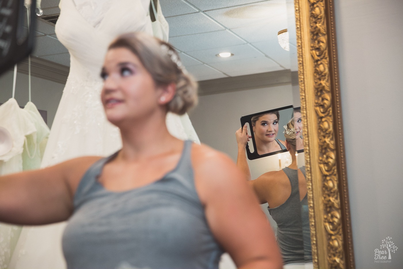 Bride looking in mirror at her hair with two wedding dresses hanging in the background