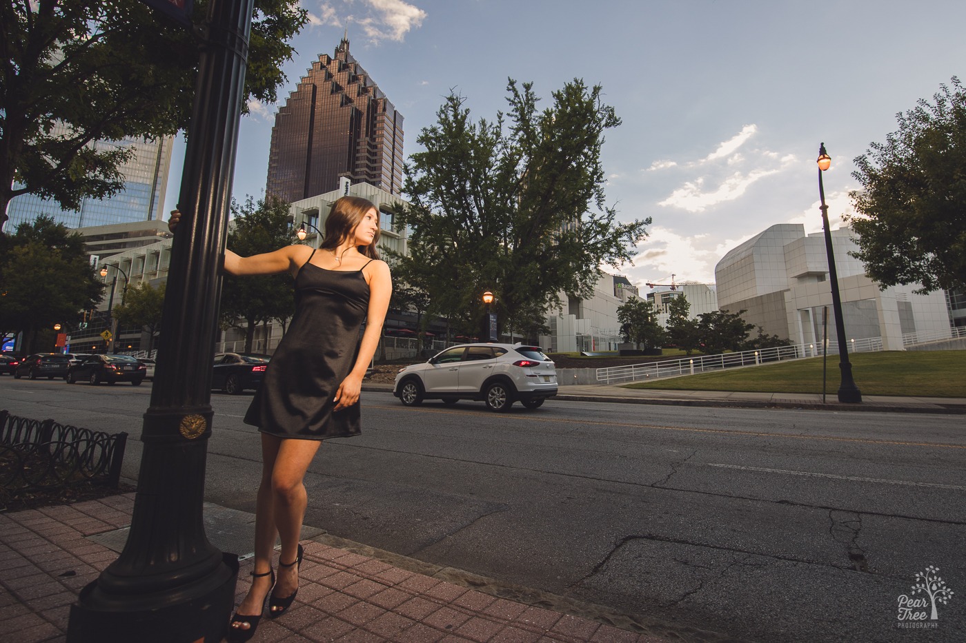 Beautiful high school senior girl in black silk dress and heels holding onto a light post in front of the High Museum