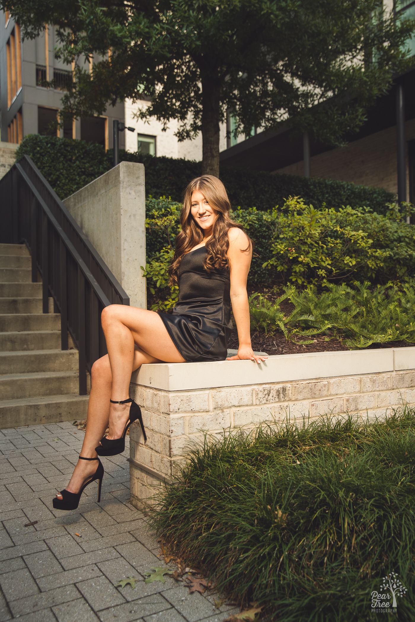 Smiling teenage girl sitting tall on a brick wall and wearing a black silk dress