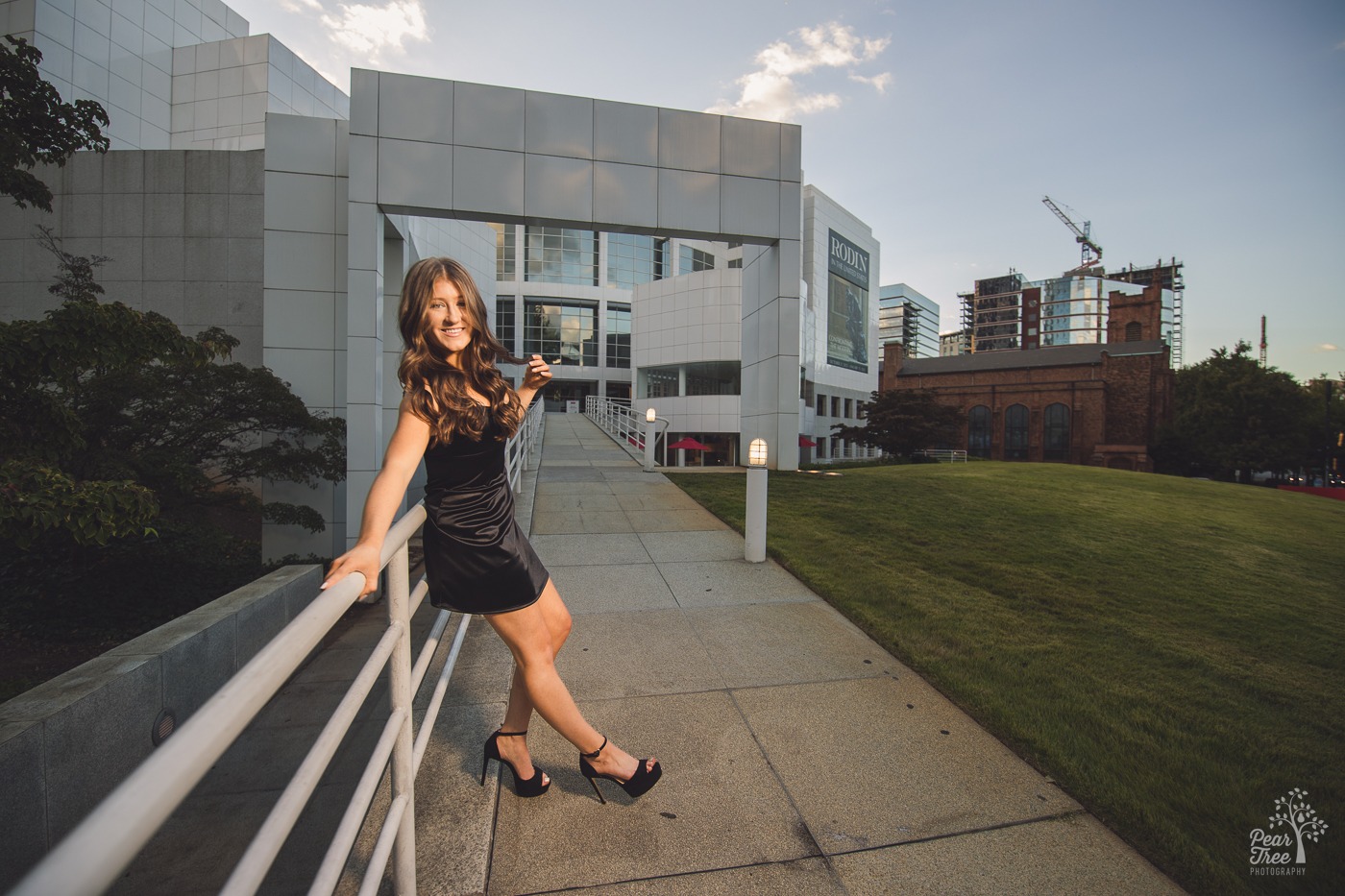 Pretty teenage girl in black silk dress and high heels leaning on a railing in front of the High Museum of Art