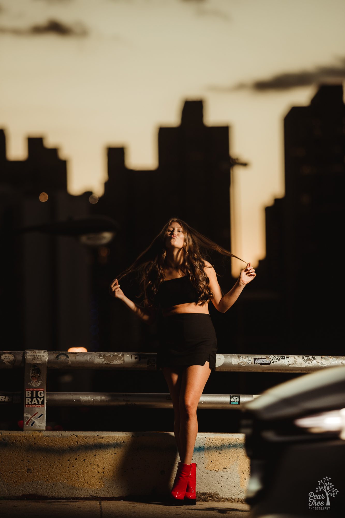 High school senior girl being silly and blowing a kiss on the Jackson Street Bridge while playing with her hair at sunset as a car starts to drive in front of her