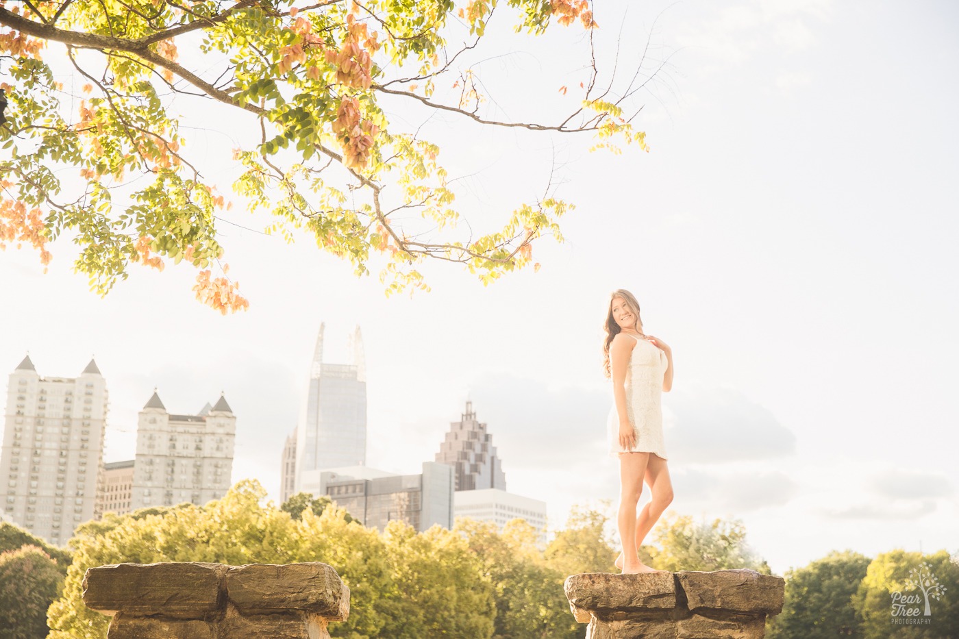 Smiling high school senior girl standing in Atlanta skyline at Piedmont Park while wearing a white lacy dress